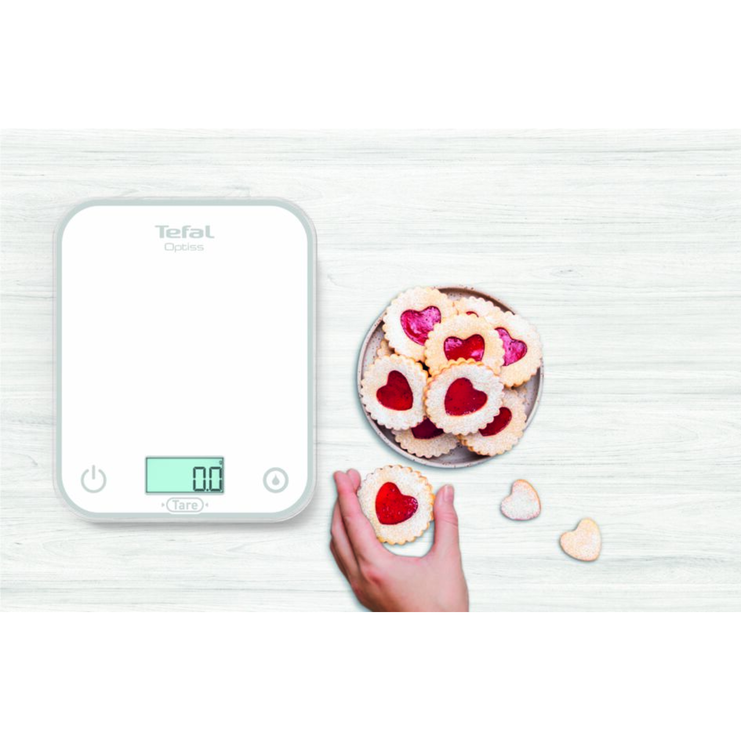 White kitchen scale with heart-shaped cookies. A hand is holding a cookie. Wooden table.