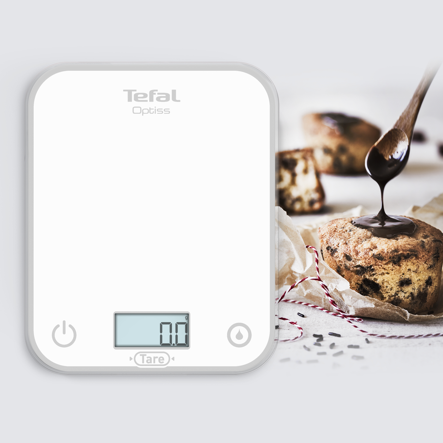 A white kitchen scale with 0.0 display. A muffin is being drizzled with chocolate from a wooden spoon.