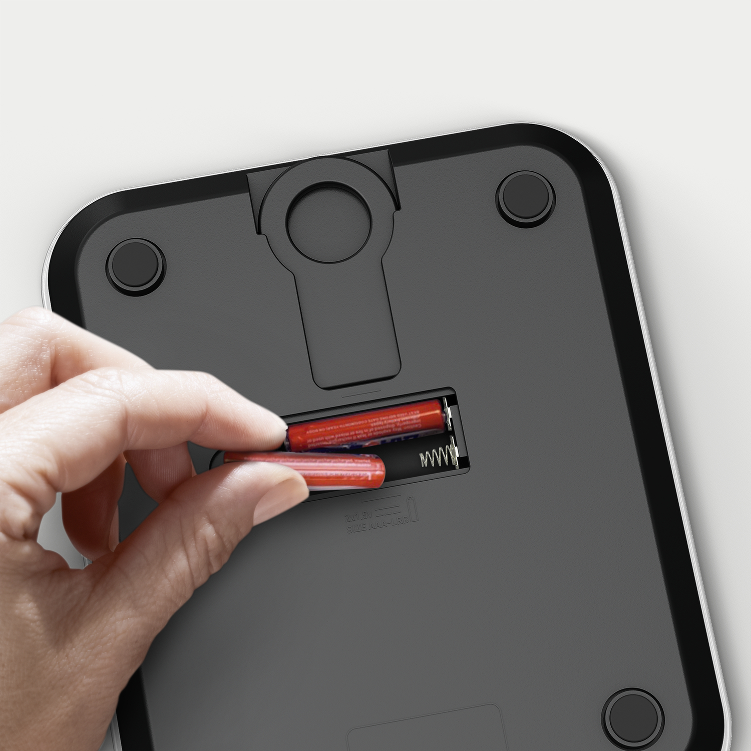 Hand inserting batteries into a scale. Red batteries, white scale with black bottom.