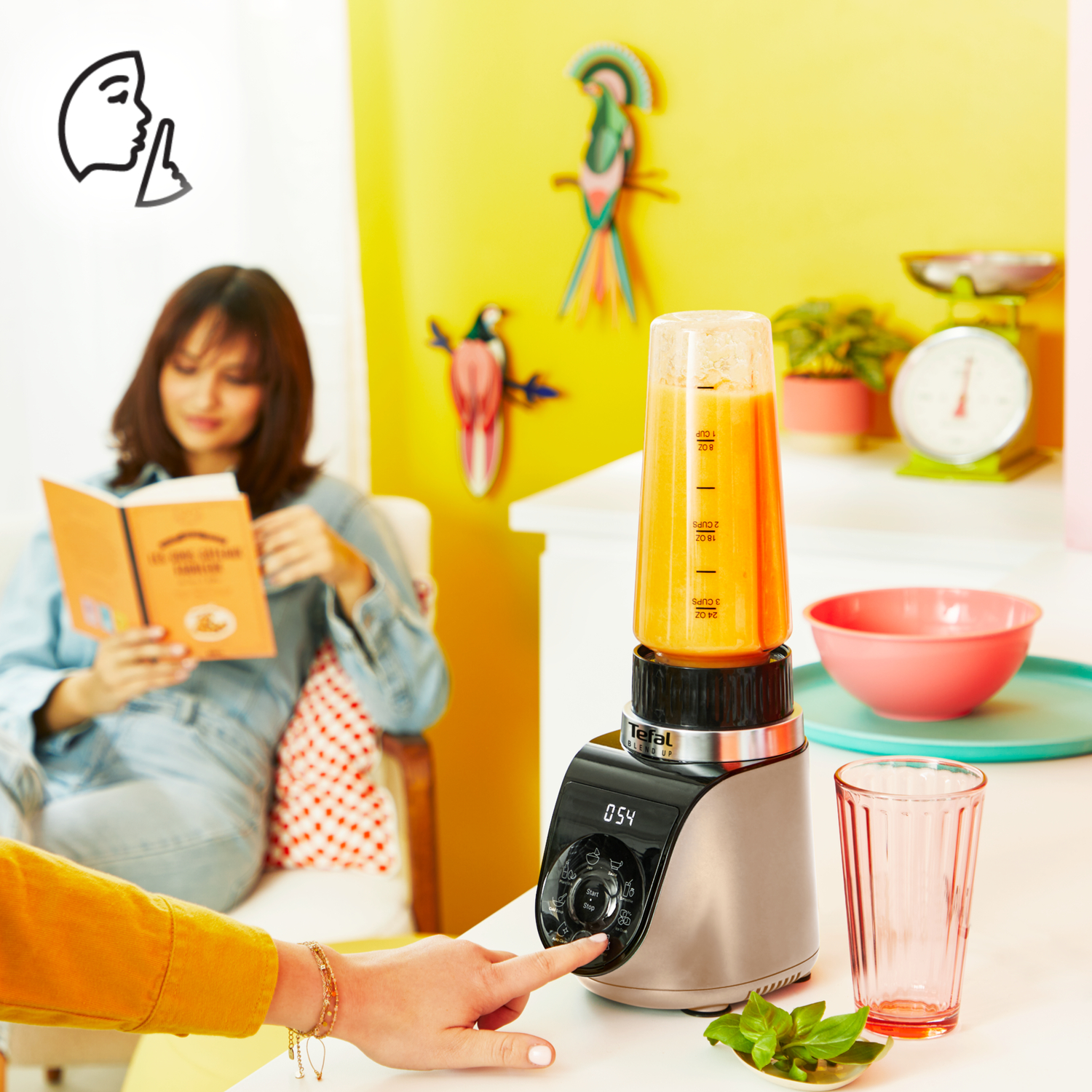 Woman using a blender. Blender is on a counter with a glass and basil. A book and parrot decor in the back.