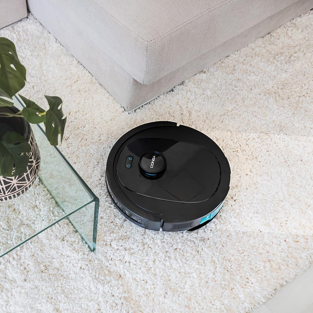 Egy fekete robotporszívó egy bolyhos fehér szőnyegen, egy üvegasztal és egy kanapé közelében.