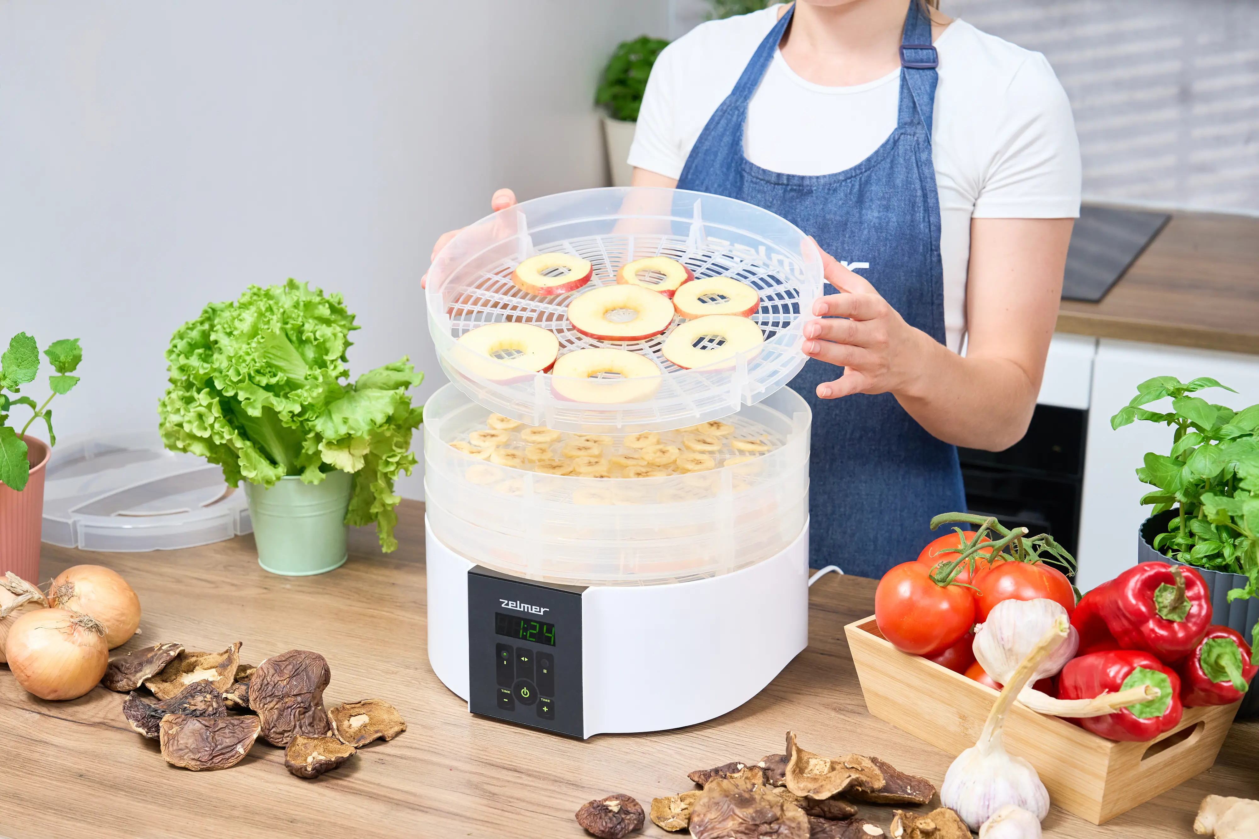 Kobieta wkłada tacę z pokrojonymi jabłkami do suszarki. Widoczne są warzywa i owoce.