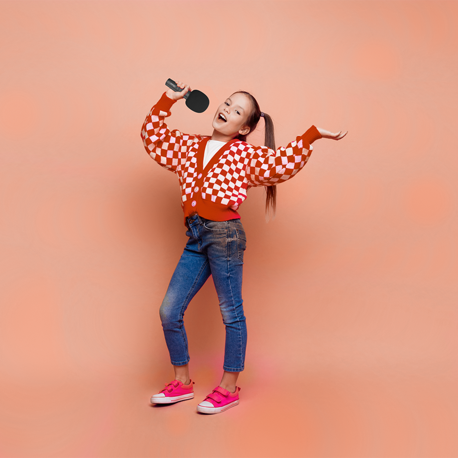 A girl sings with a ping pong paddle microphone, wearing a red checkered sweater, blue jeans, and pink shoes.