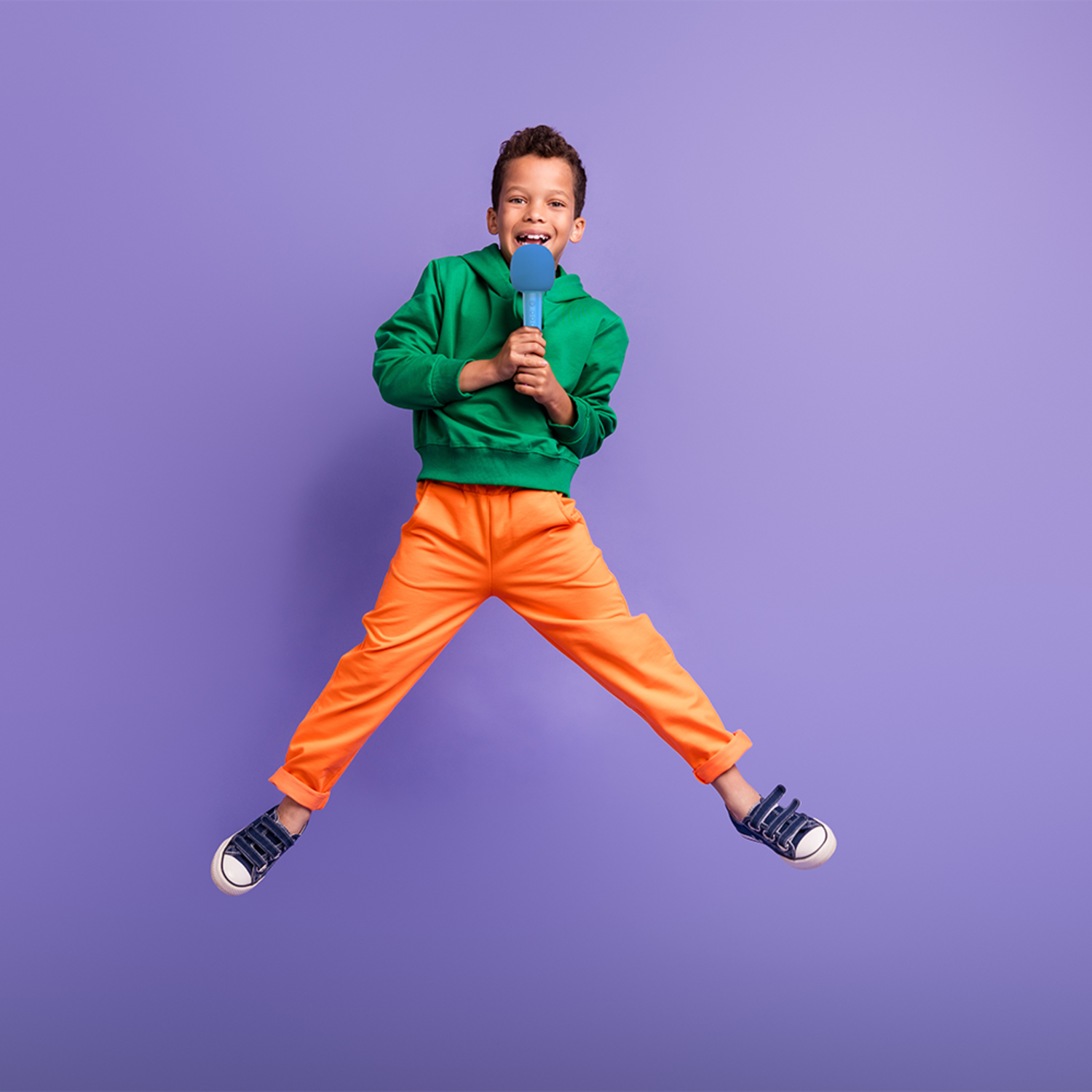 Een jongen springt in de lucht, houdt een blauwe microfoon vast, lacht, met een oranje broek en een groene hoodie.