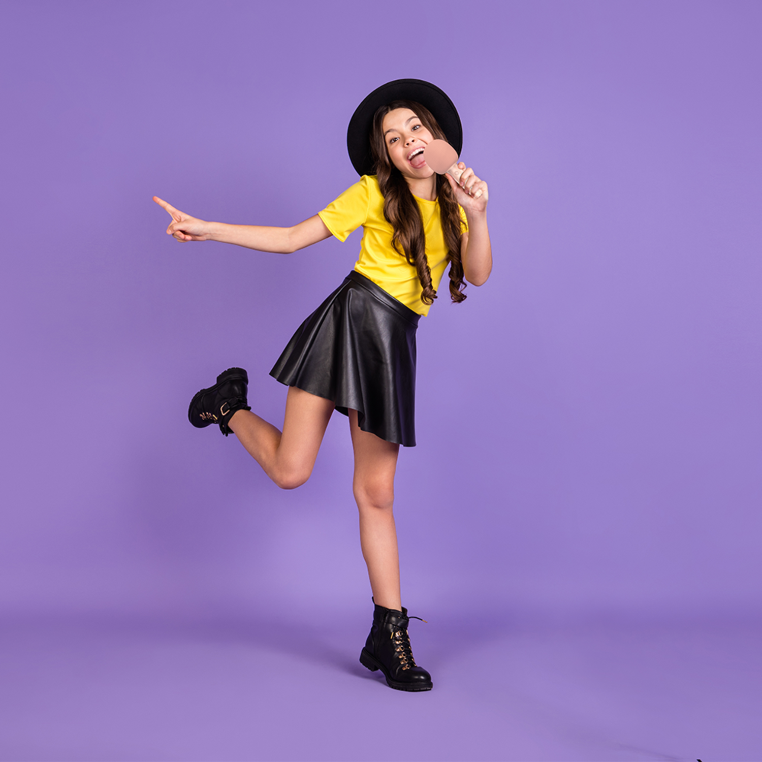 Young girl in a black hat, skirt, and yellow shirt, posing with a pink object.