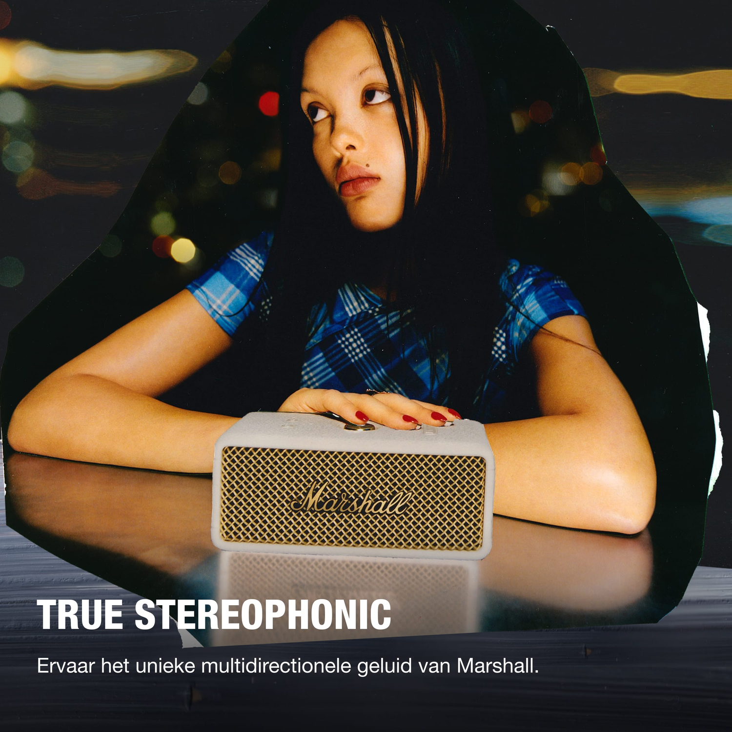 Woman with a Marshall speaker. The speaker is on a table. The background is blurred with city lights.