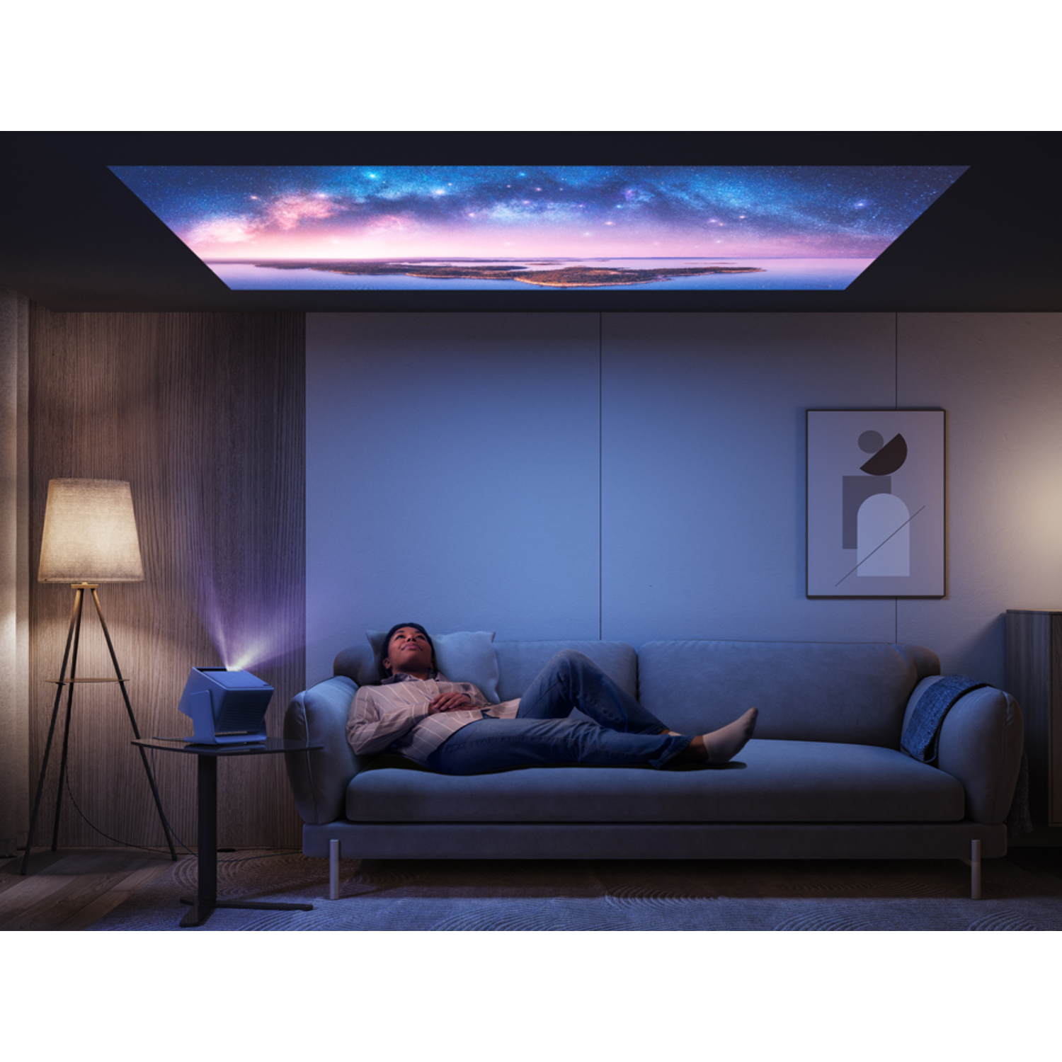 A person lying on a gray sofa watches a starry sky projected on the ceiling. A lamp and a projector are also present.