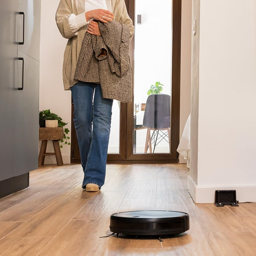 Nő a szobában, a padlón robotporszívóval. Kabátot tart.