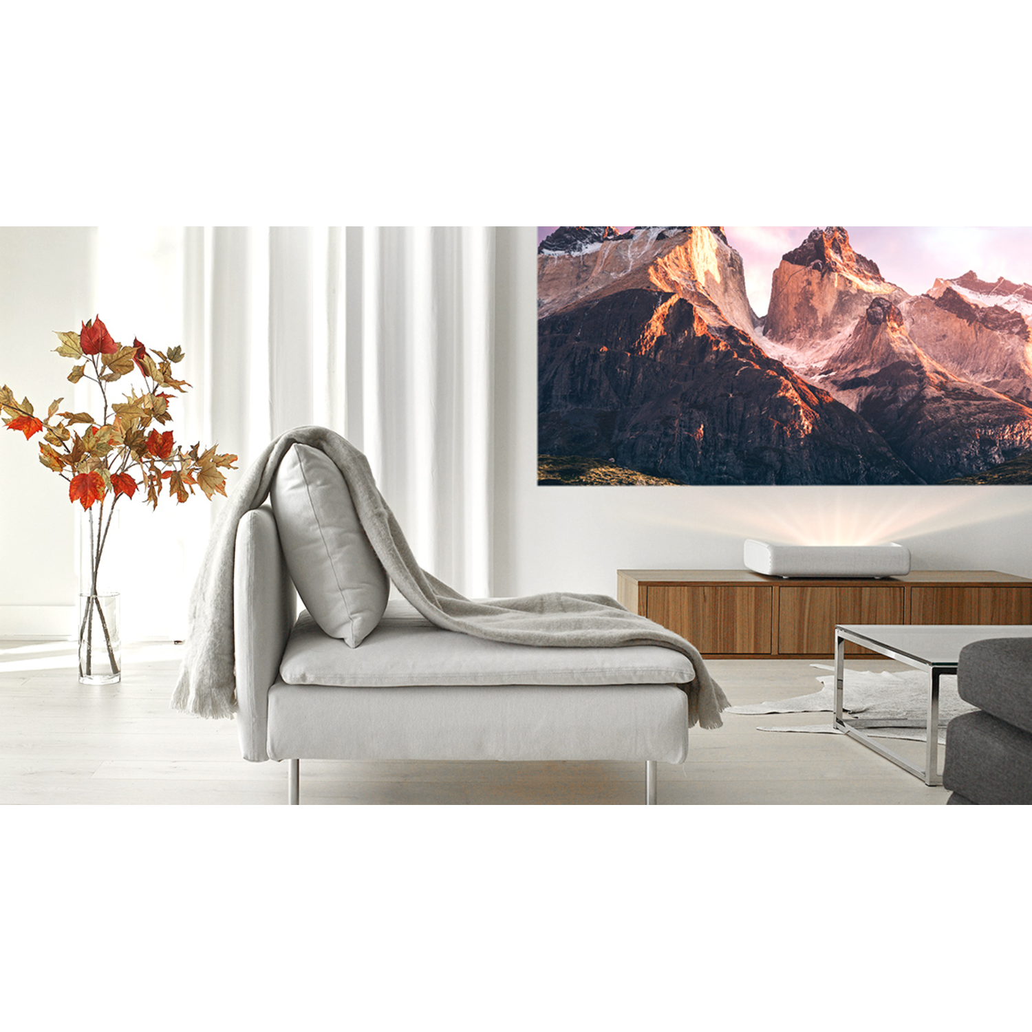 Een projector toont een berglandschap op een witte muur, met een bank en plant op de voorgrond.