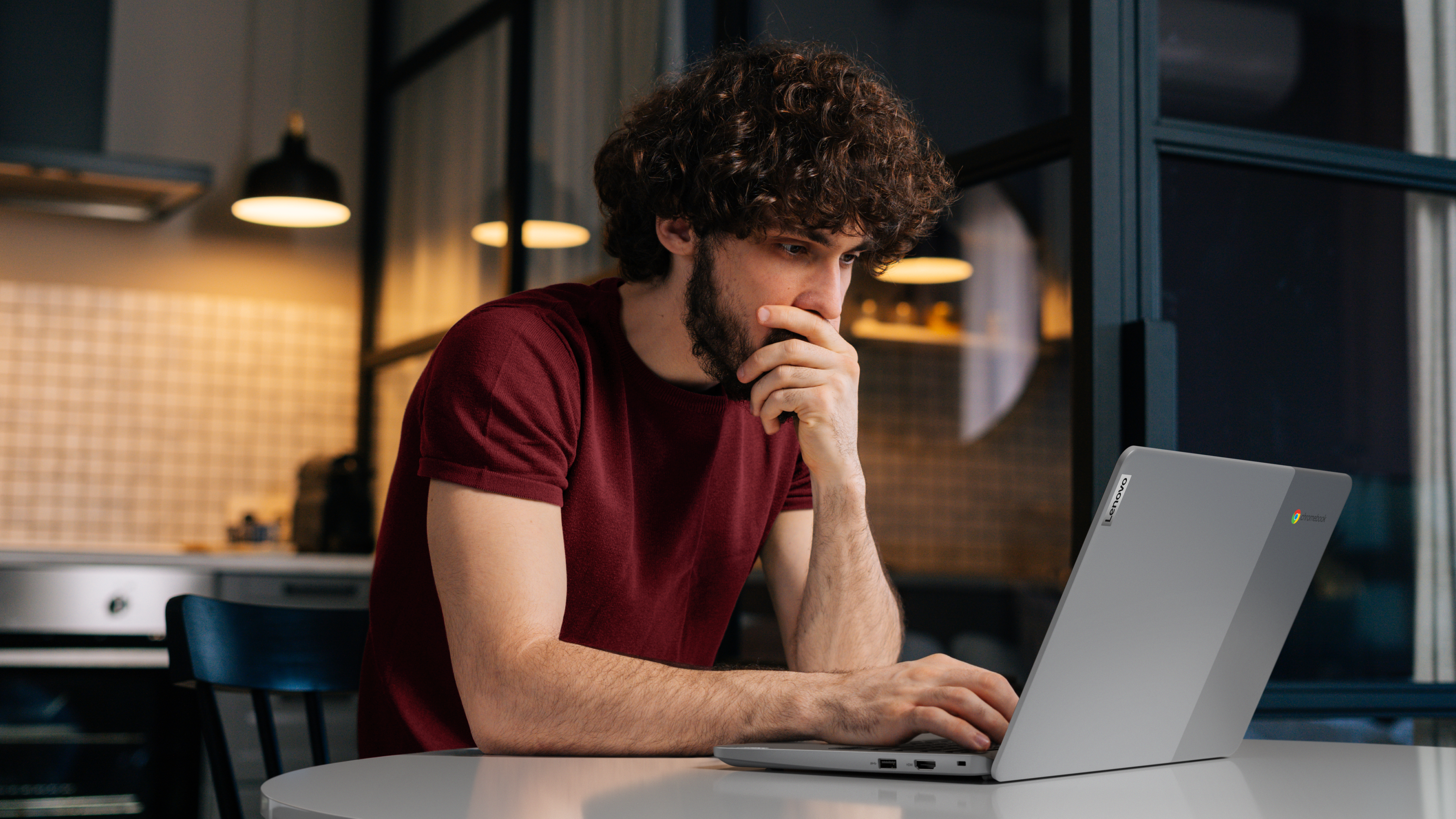 Ein Mann mit lockigen Haaren schaut auf einen Laptop. Er ist in einem Raum mit einem Tisch und einer Küche.