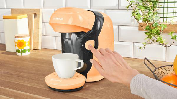 Die Hand einer Person drückt eine Taste an einer orangefarbenen und schwarzen Kaffeemaschine neben einer Tasse.