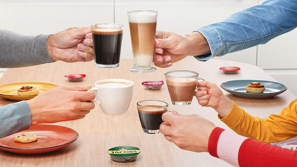 Hände halten verschiedene Kaffeetassen und Kapseln auf einem Holztisch. Kekse und Zimtschnecken sind vorhanden.