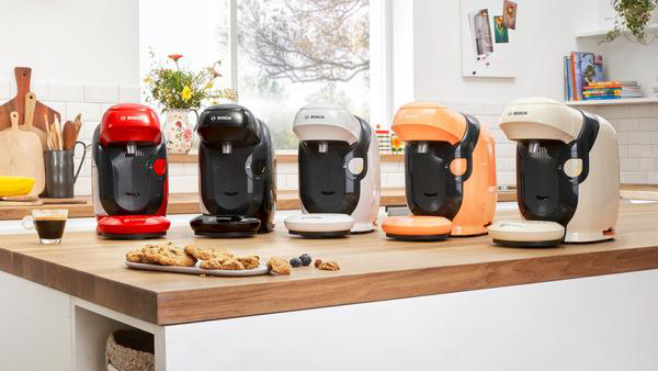 Fünf verschiedenfarbige Kaffeemaschinen auf einer Holz-Arbeitsplatte mit Keksen und einer Tasse Kaffee.