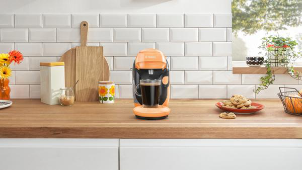 Eine orangefarbene Kaffeemaschine mit einer Tasse, Keksen auf einem Teller und Blumen auf einer Holztheke.