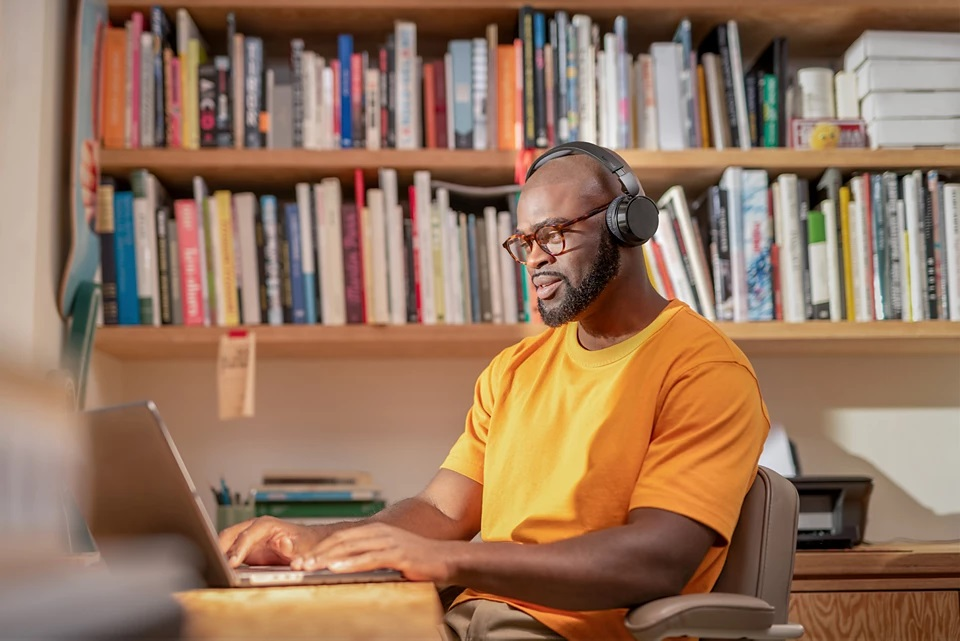 Czarny mężczyzna ze słuchawkami i okularami korzystający z laptopa w domowym biurze.
