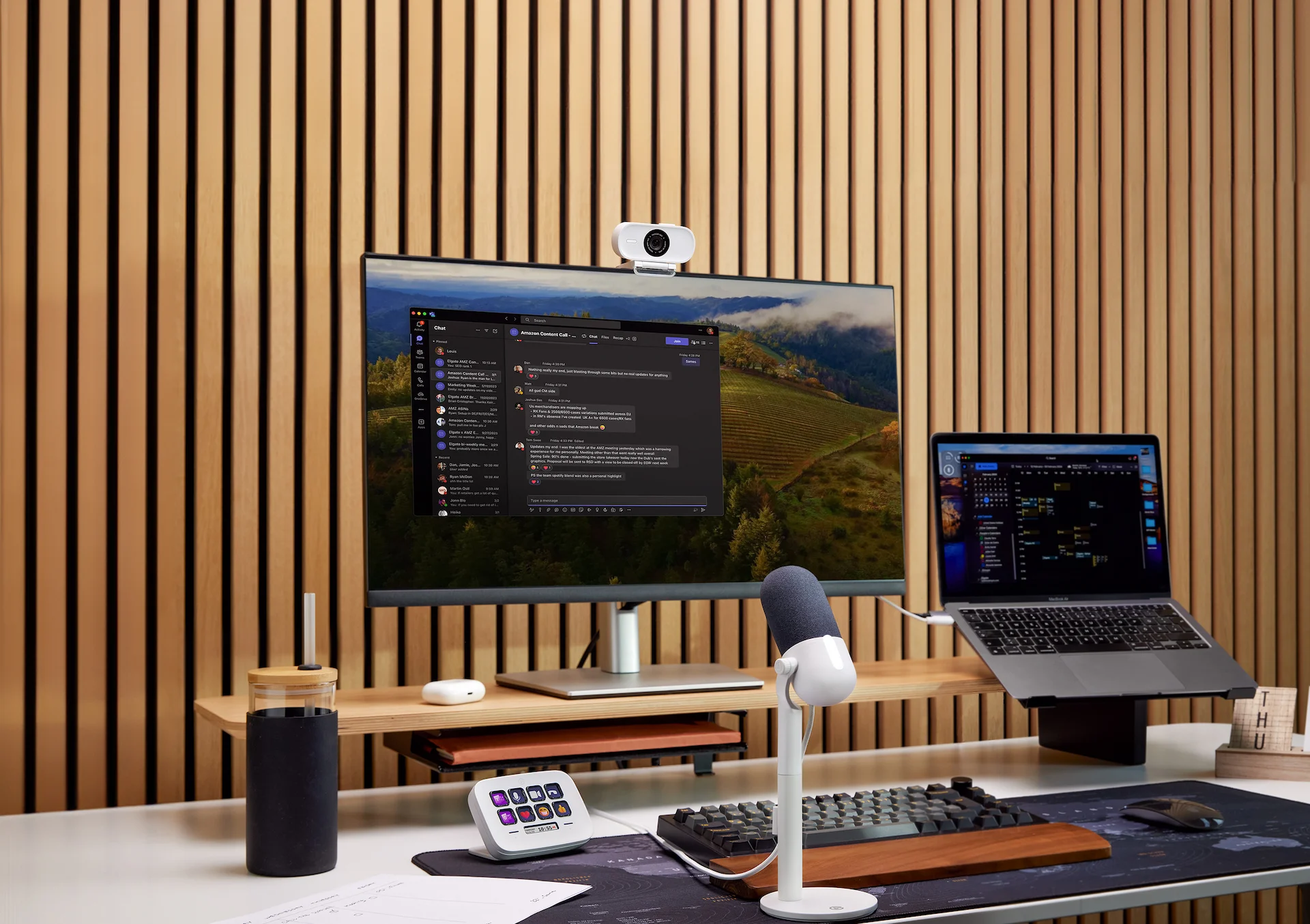 Ein moderner Arbeitsplatz mit Monitor, Laptop, Mikrofon, Tastatur und anderem Zubehör auf einem Holzschreibtisch.