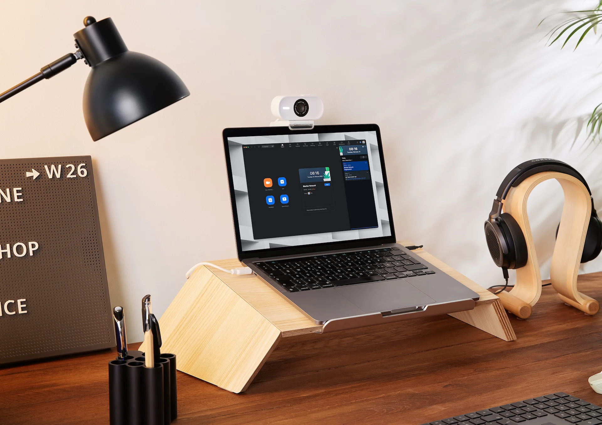 Laptop auf Holzständer mit Webcam, Kopfhörer, Schreibtischlampe und Stiften auf Holzschreibtisch. Zoom-Meeting auf dem Bildschirm.