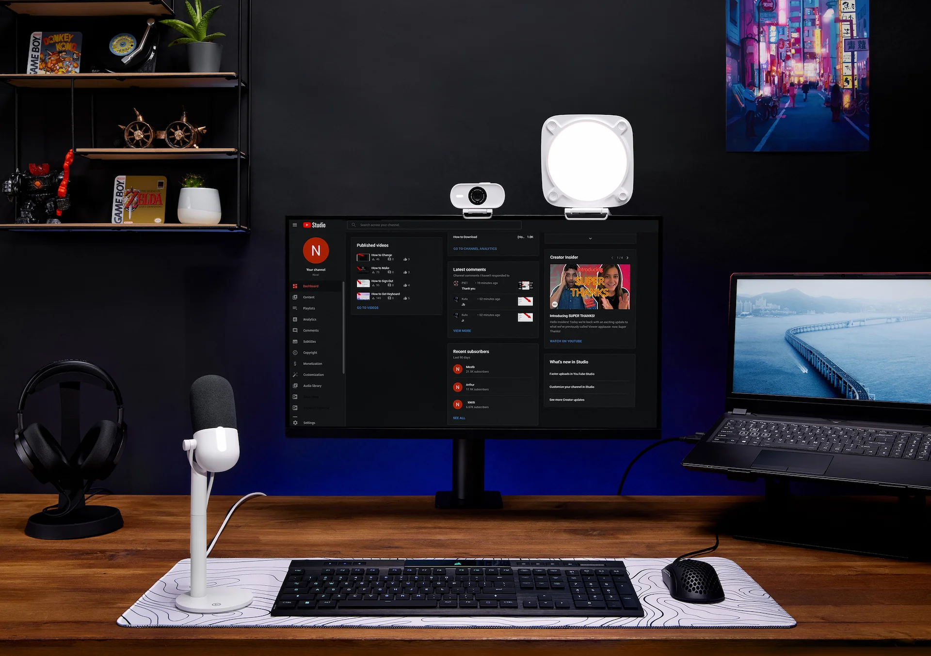 Ein Schreibtisch mit Monitor, Tastatur, Mikrofon und Laptop. Ein Regal mit Dekoration im Hintergrund. Ein Stadtkunstwerk.
