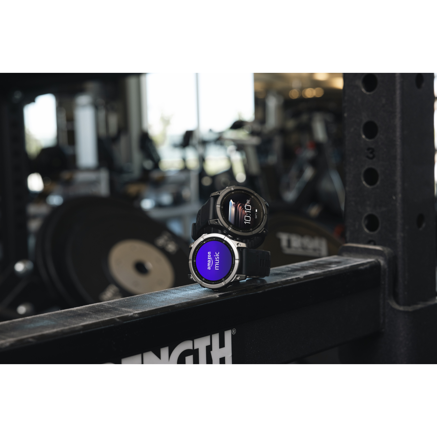 Deux smartwatches sur un rack métallique dans une salle de sport. Logo Amazon Music.