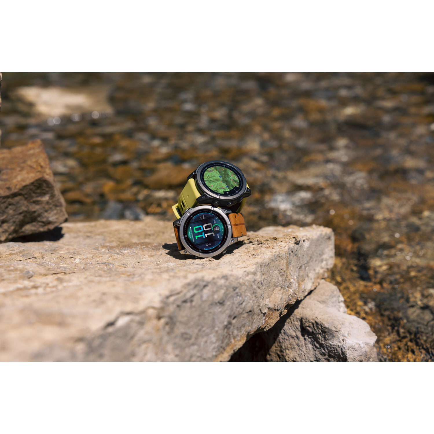 Twee smartwatches, een met een gele band, de andere met een bruine, op een steen bij water.