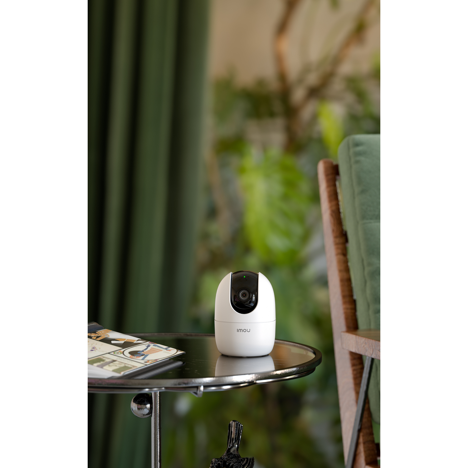 A white security camera on a table next to a magazine, with green plants in the background.