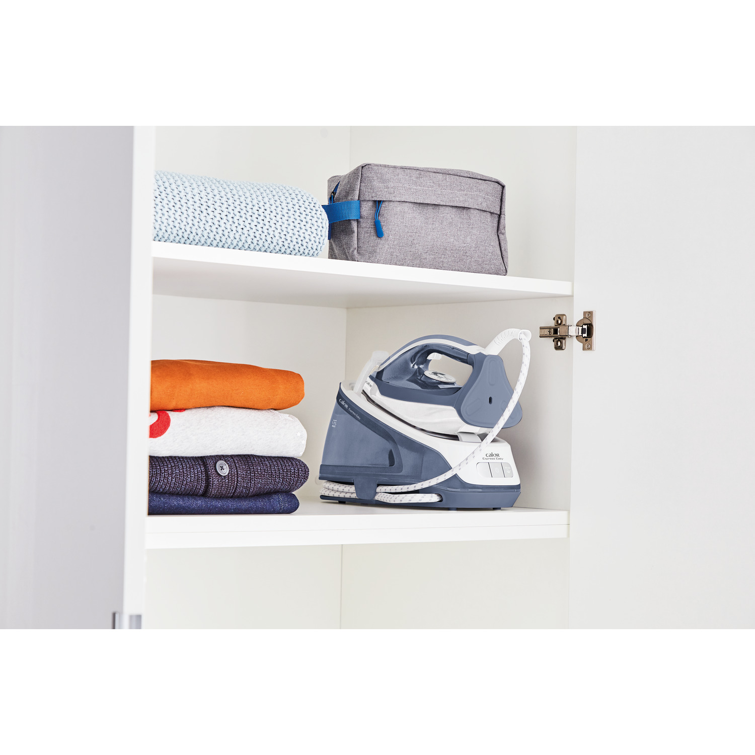 Open white cabinet with iron and clothes on shelves. Gray bag on top shelf.