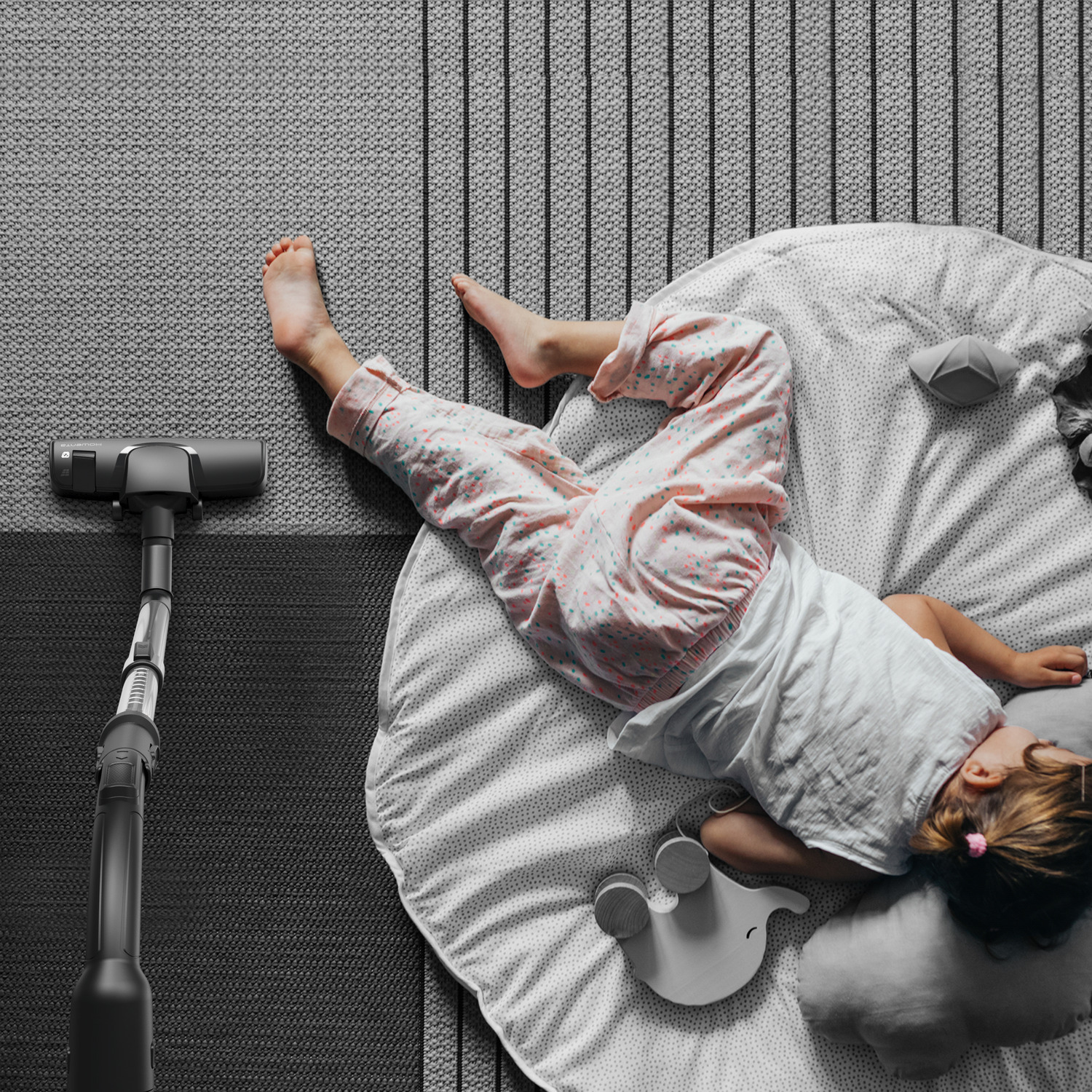 A child lies on a mat with a vacuum cleaner nearby. The child wears pink pants.