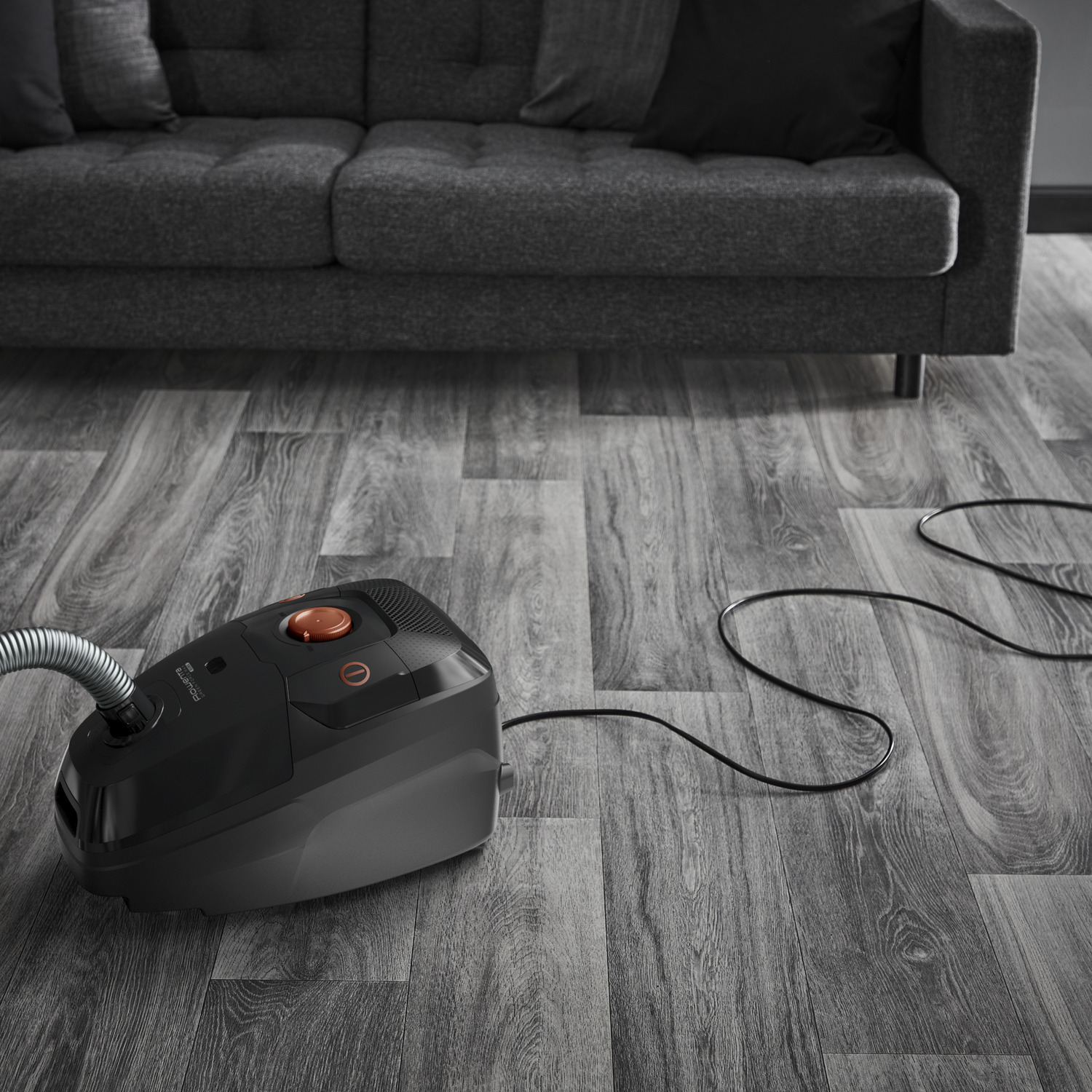 Black vacuum cleaner on a grey wooden floor near a grey couch. Black cable.
