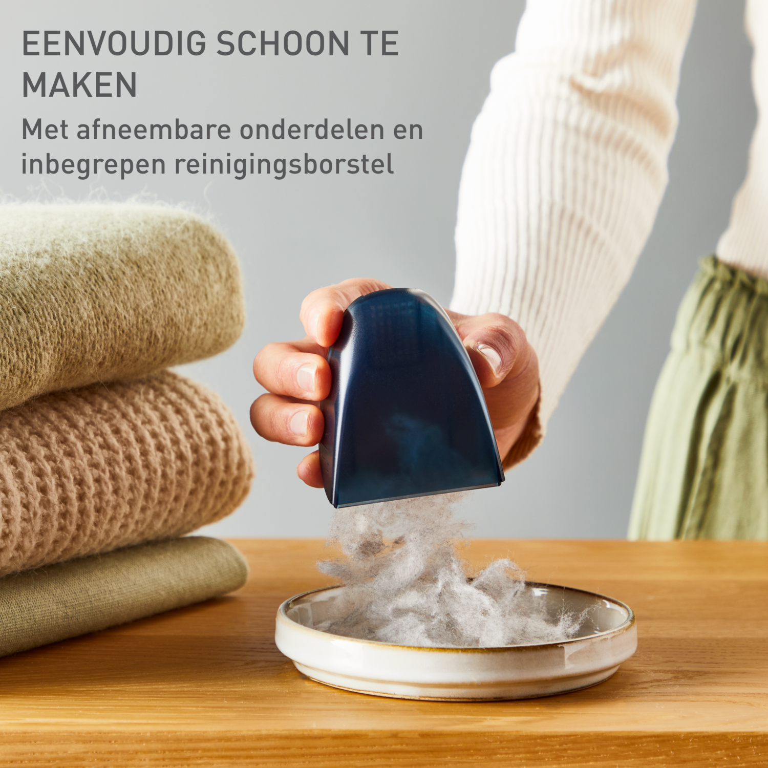 Hand die een pluizenverwijderaar boven een kom met pluisjes houdt, naast stapels gevouwen kleding.