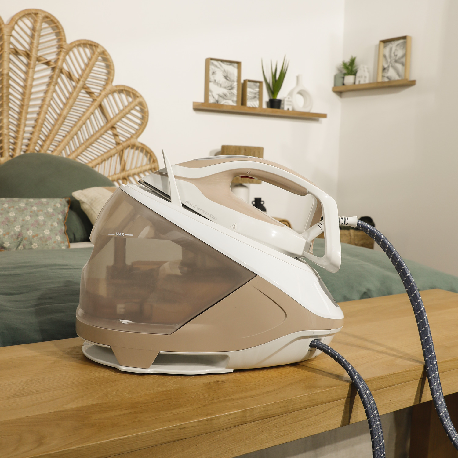 A beige and white iron on a wooden table, with a bed and decor in the background.