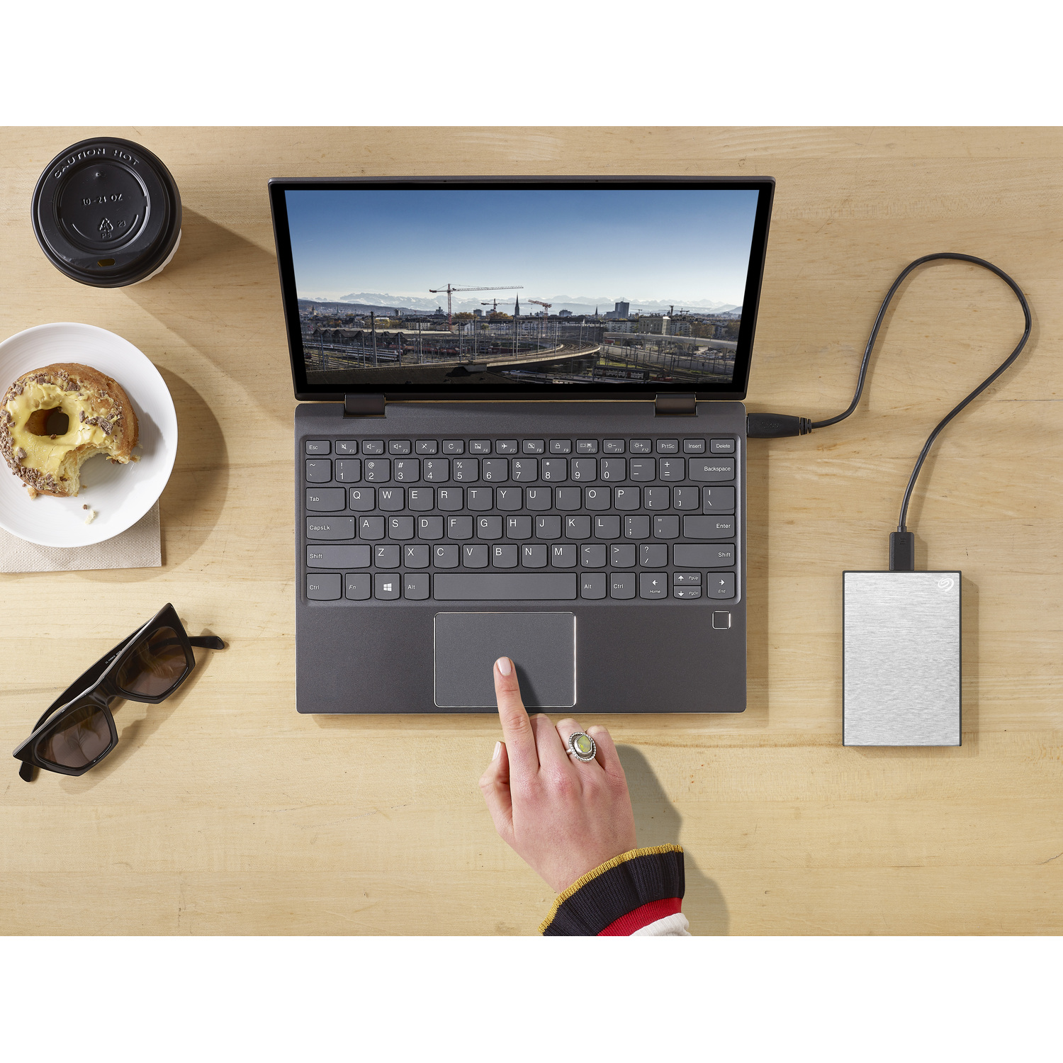 Ordinateur portable ouvert sur un bureau avec disque dur externe, lunettes, café et donut. Une main touche le pavé tactile.