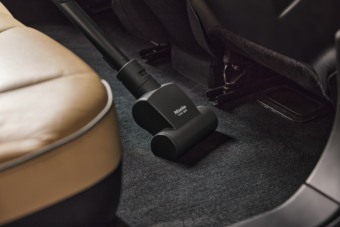Schwarzer Staubsaugerkopf reinigt den grauen Teppich eines Autos. Beiger Sitz im Hintergrund.