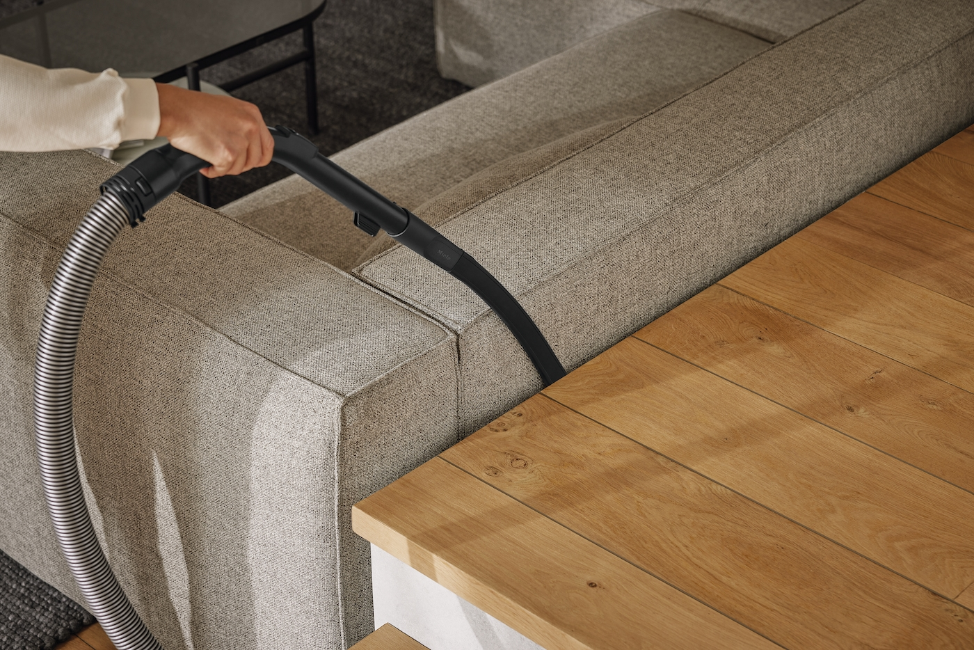 Person saugt hellgraues Sofa mit dunklem Staubsauger. Ein Holztisch steht neben dem Sofa.