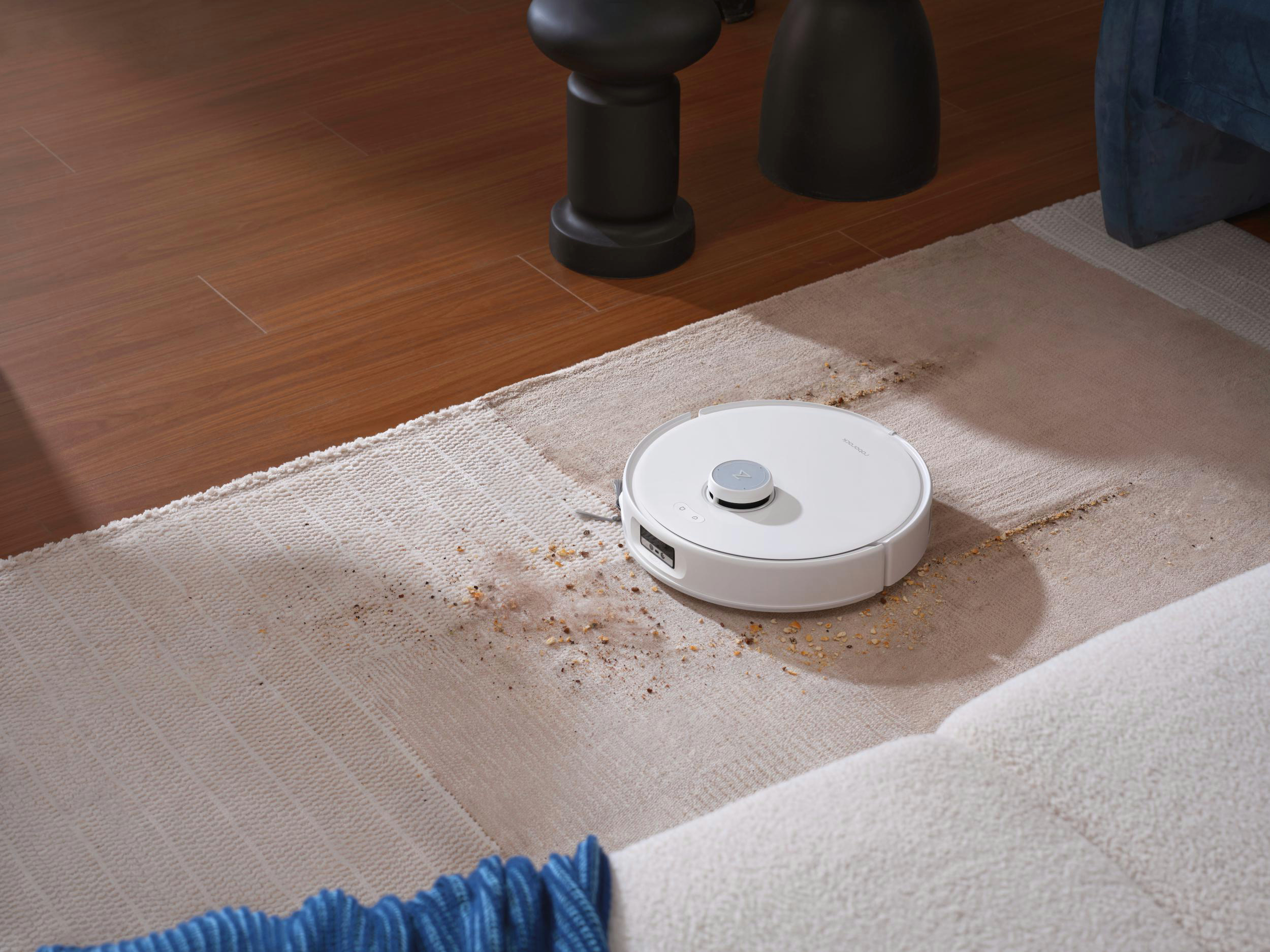 Roboter-Staubsauger auf einem Teppich, der reinigt. Braune Rückstände sind sichtbar. Teil eines Sofas ist zu sehen.
