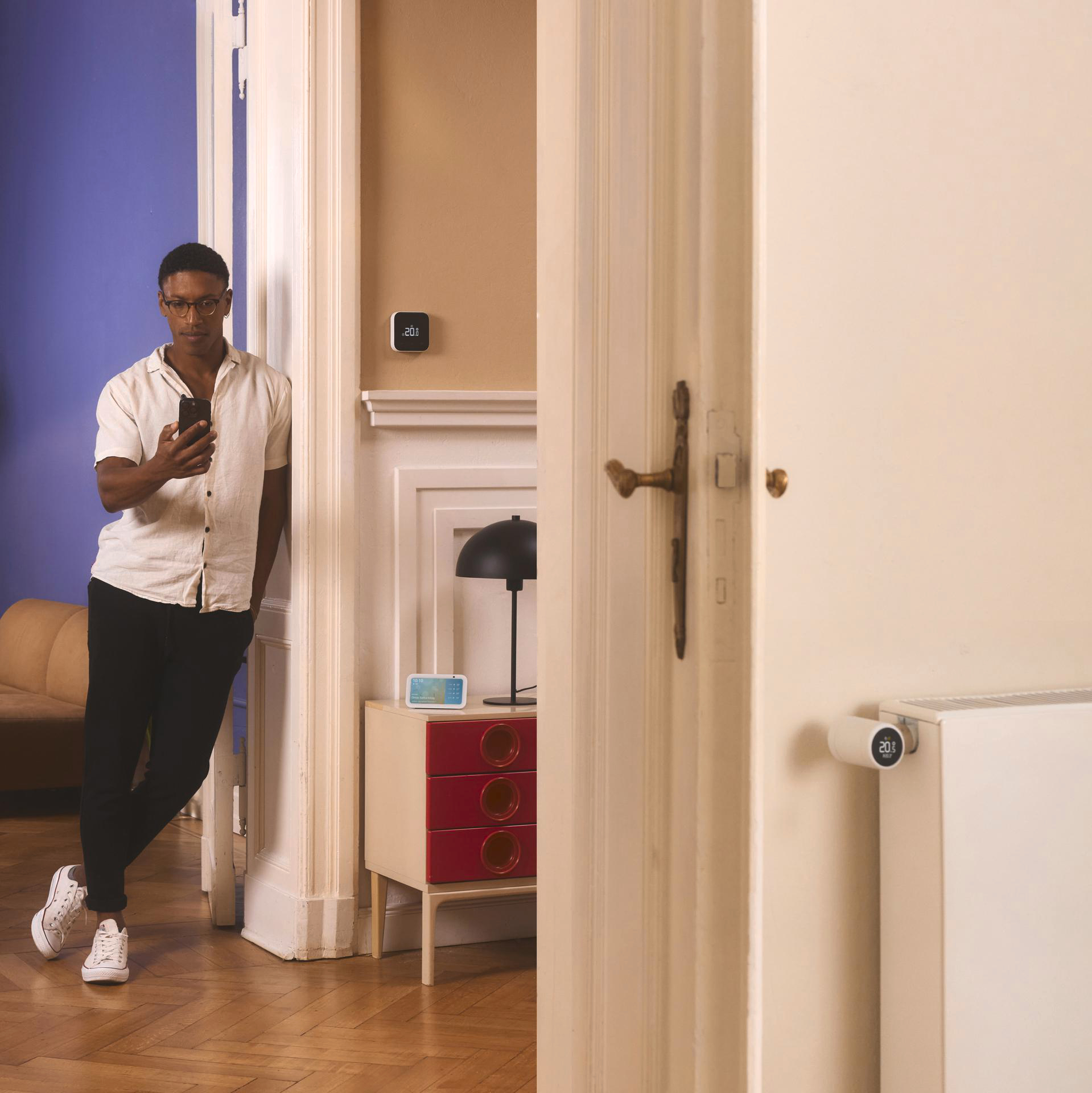 Mann in einem Raum, der ein Telefon hält. Ein intelligenter Thermostat hängt an der Wand. Beigefarbene Wand, weißer Türrahmen.