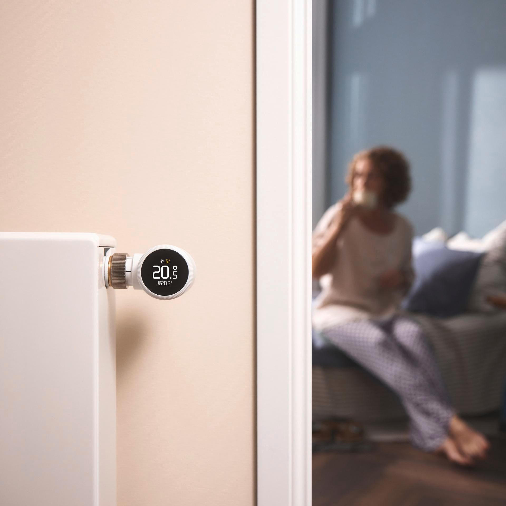 Smart-Thermostat an einer Wand. Eine Frau sitzt im verschwommenen Hintergrund auf einem Sofa.