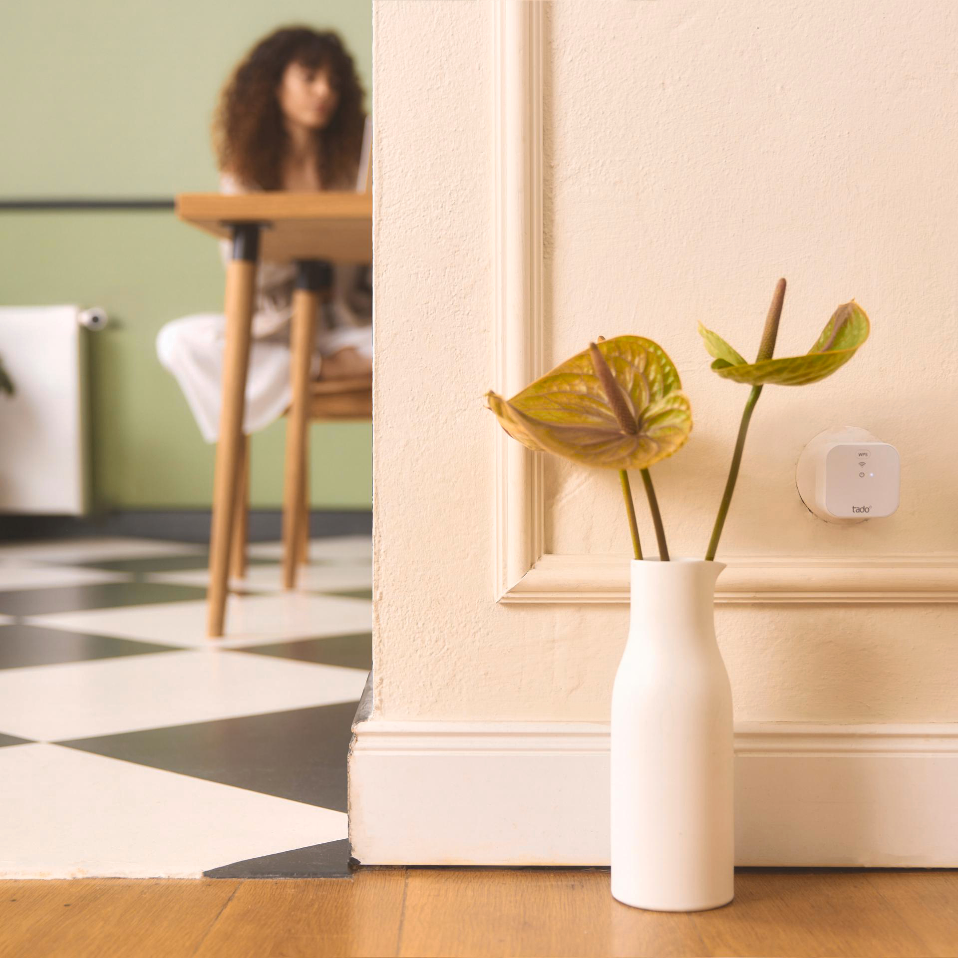 Innenraum: eine weiße Vase mit Blumen, eine Frau und ein Smart-Home-Gerät an der Wand.