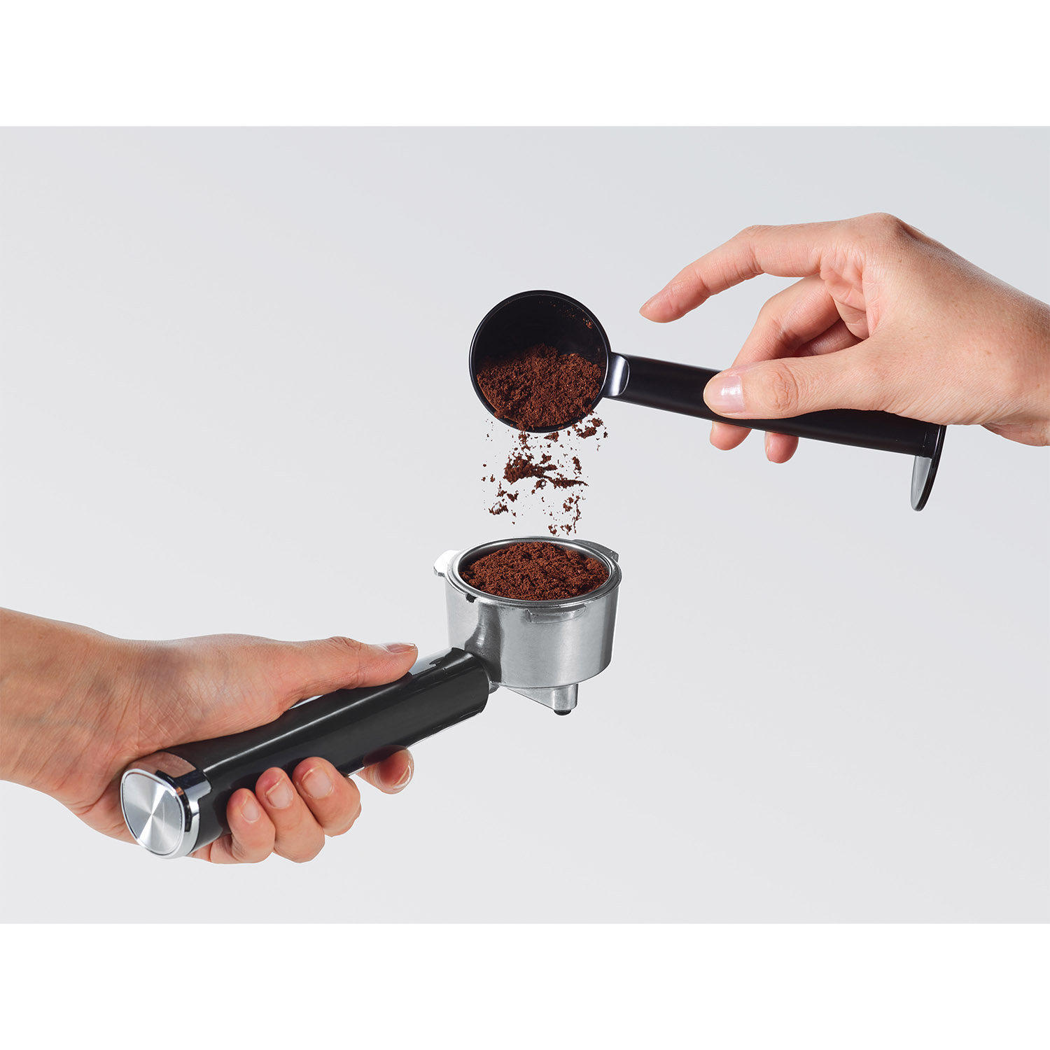 Mani che riempiono un portafiltro con caffè macinato da un cucchiaio su sfondo bianco.