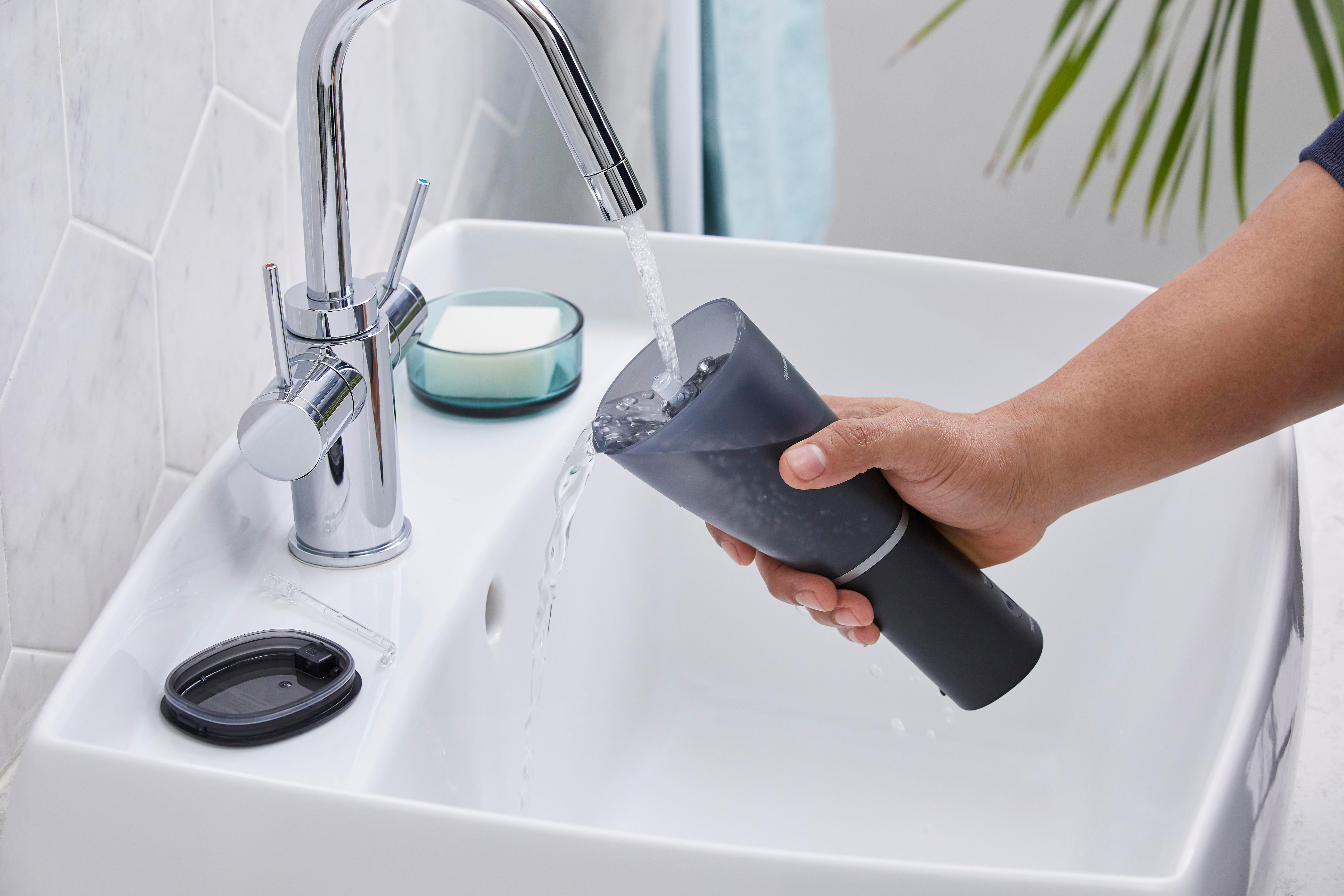 Eine Hand gießt Wasser aus einem dunkelgrauen Behälter in ein weißes Waschbecken.