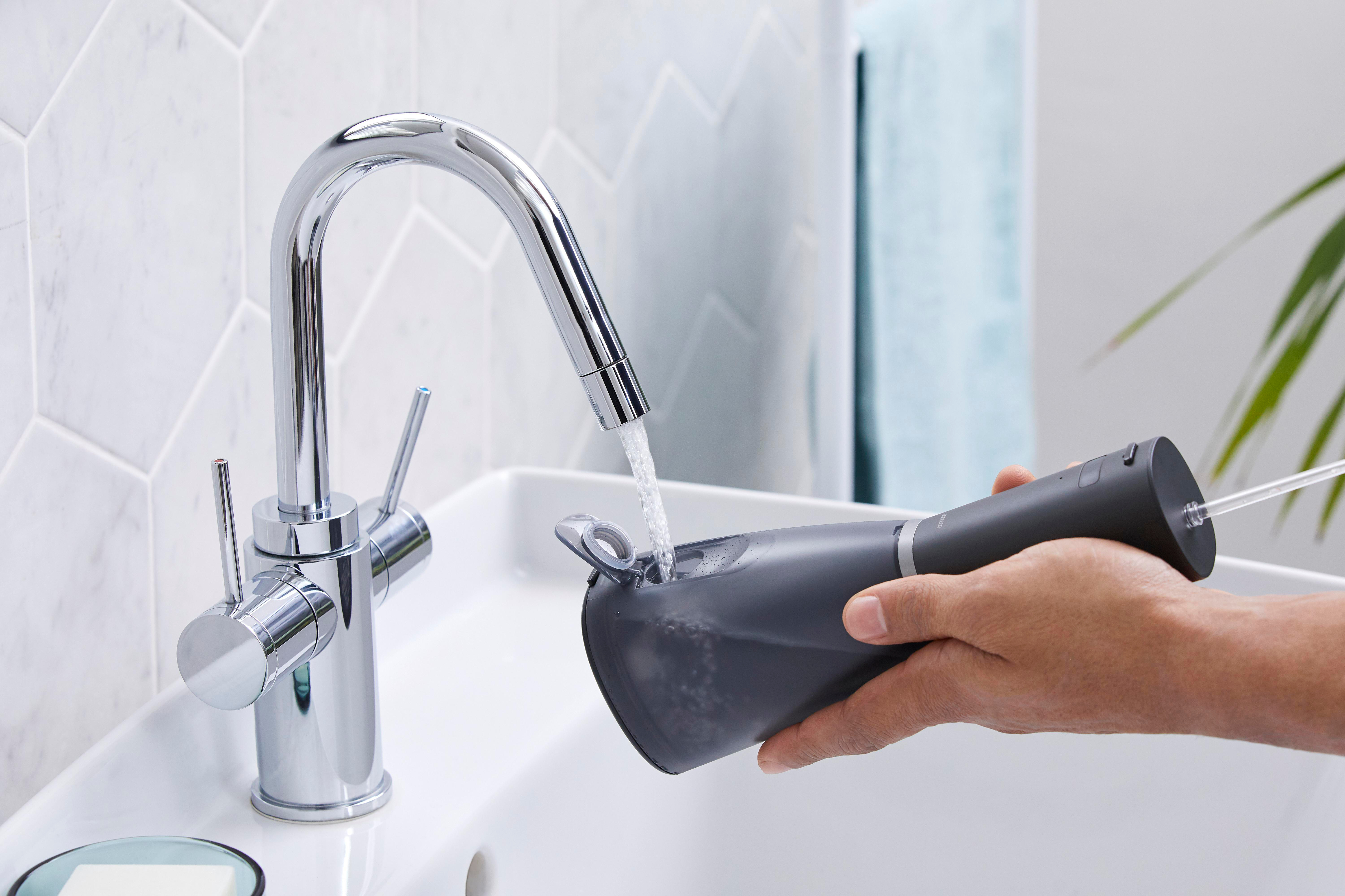 Hand befüllt einen schwarzen Zahn-Irrigator von einem Chrom Wasserhahn. Weißes Waschbecken sichtbar.