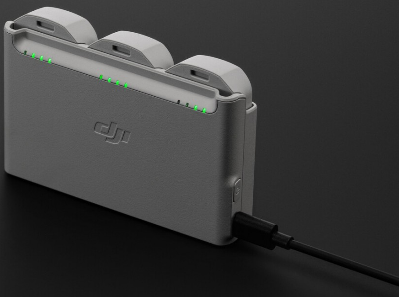 Eine graue DJI-Ladestation mit drei Steckplätzen, grünen Lichtern und einem eingesteckten schwarzen Ladekabel.