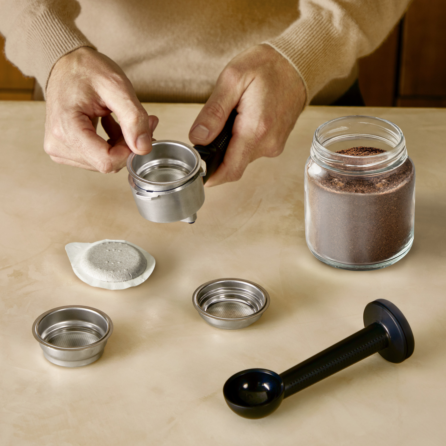 Hands preparing to make coffee, with a portafilter, filters, coffee grounds, and a scoop.