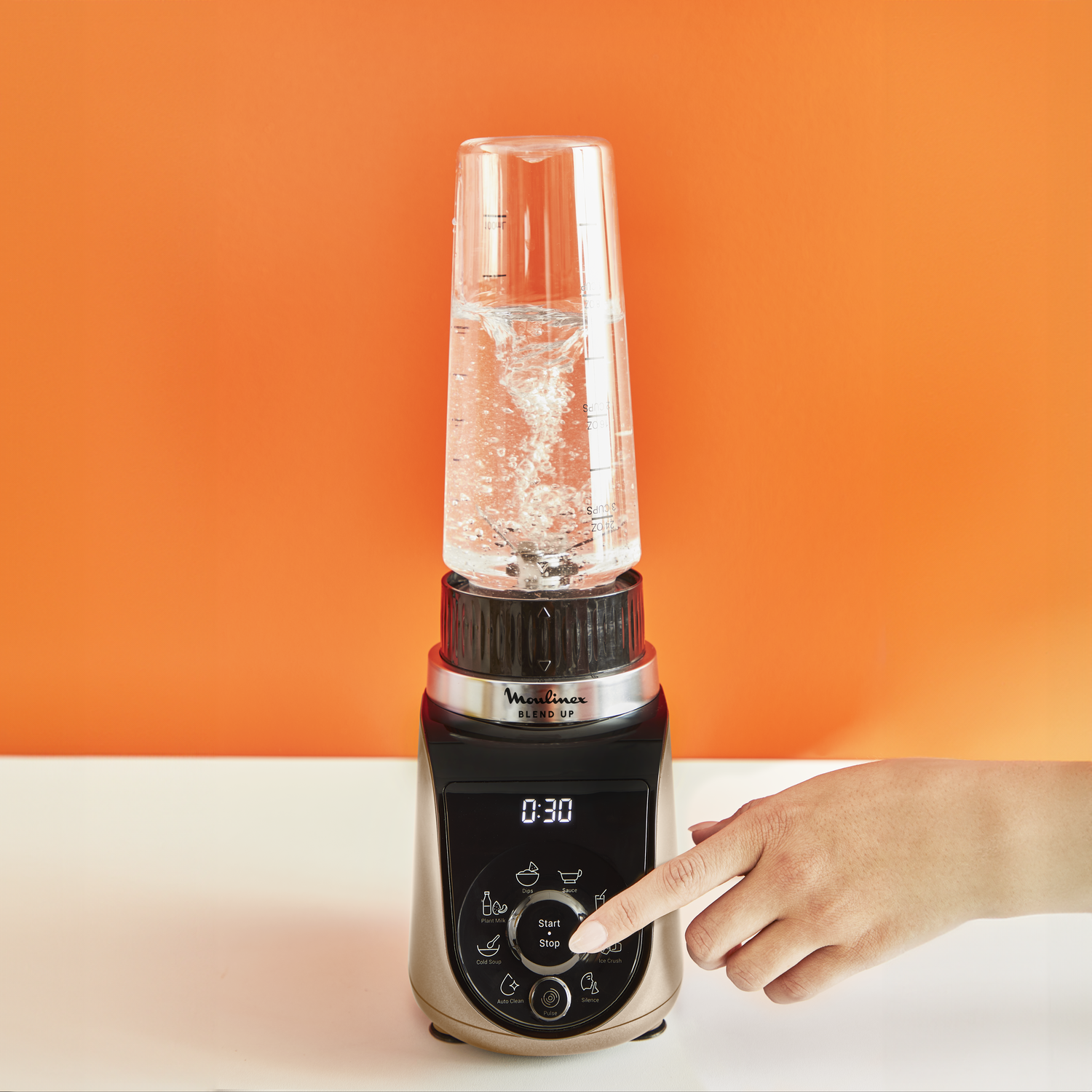 Une main appuie sur un bouton d'un blender or et noir avec de l'eau à l'intérieur.