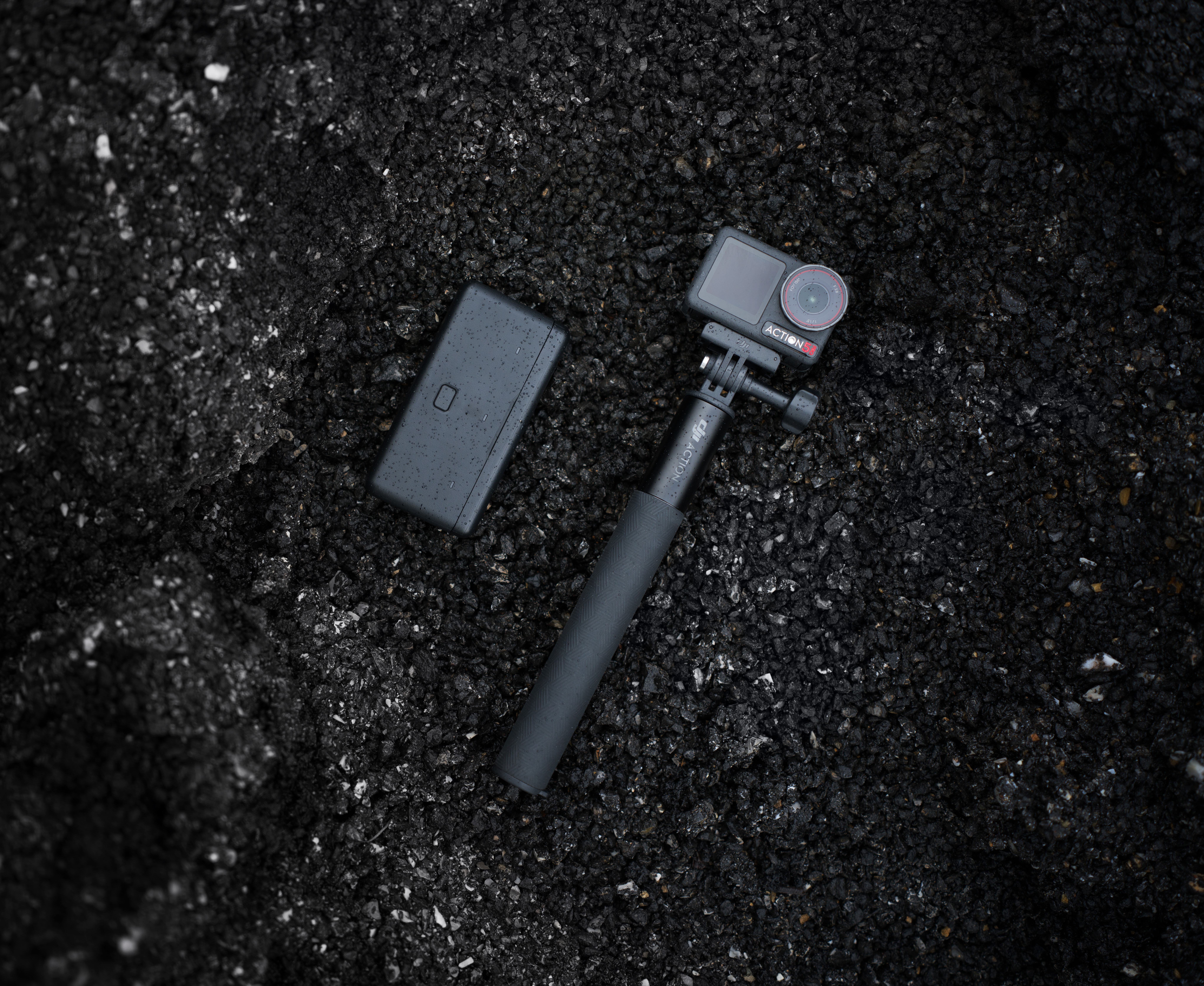 Action-Kamera, Akkupack und Selfie-Stick auf schwarzem Asphalt.