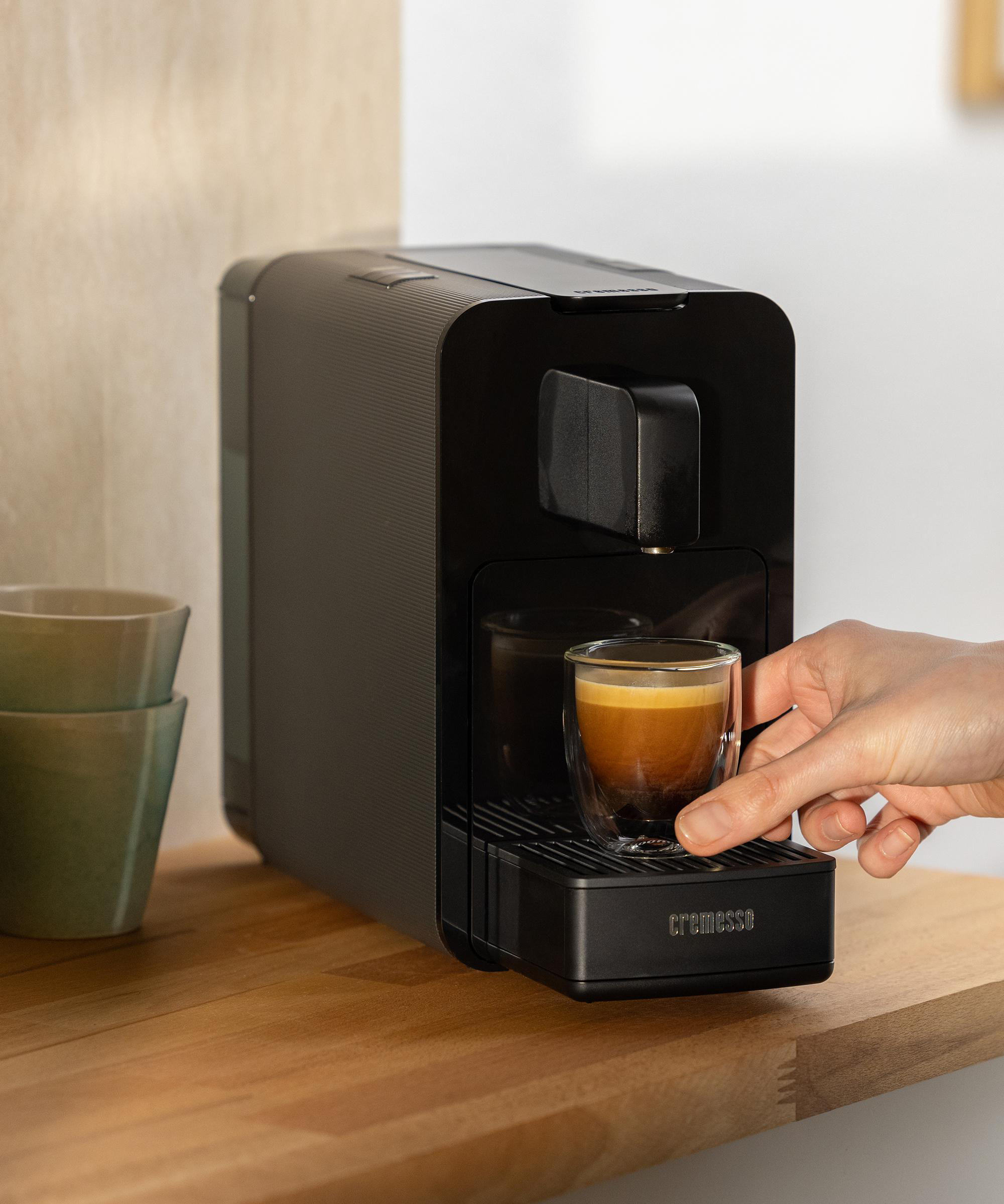 Person hält Kaffeeglas, Kaffeemaschine sagt 'cremesso', auf einem Holztisch.