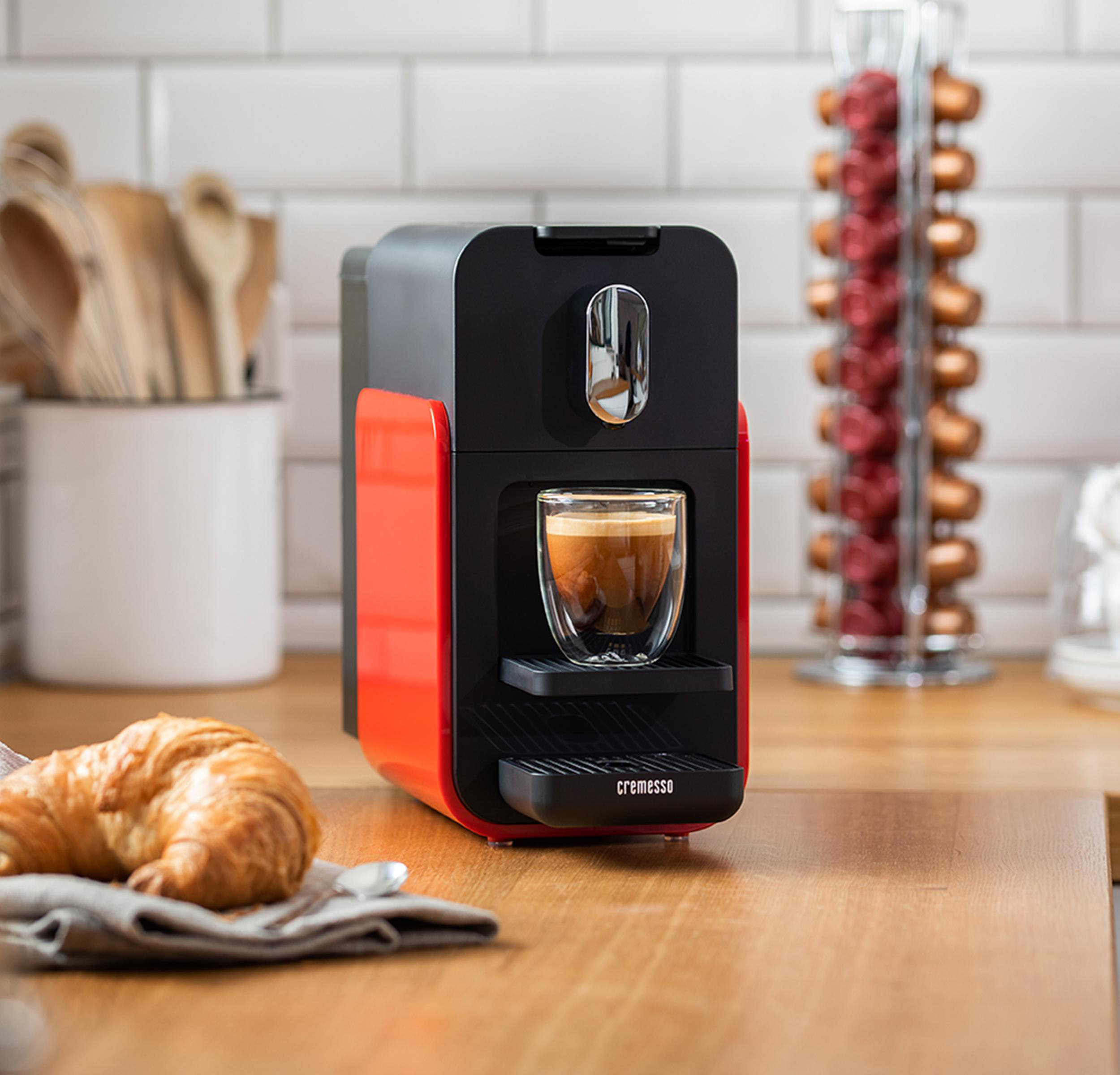 Eine rot-schwarze Kaffeemaschine mit einem Glasbecher mit Kaffee, einem Croissant und Kaffeekapseln dahinter.