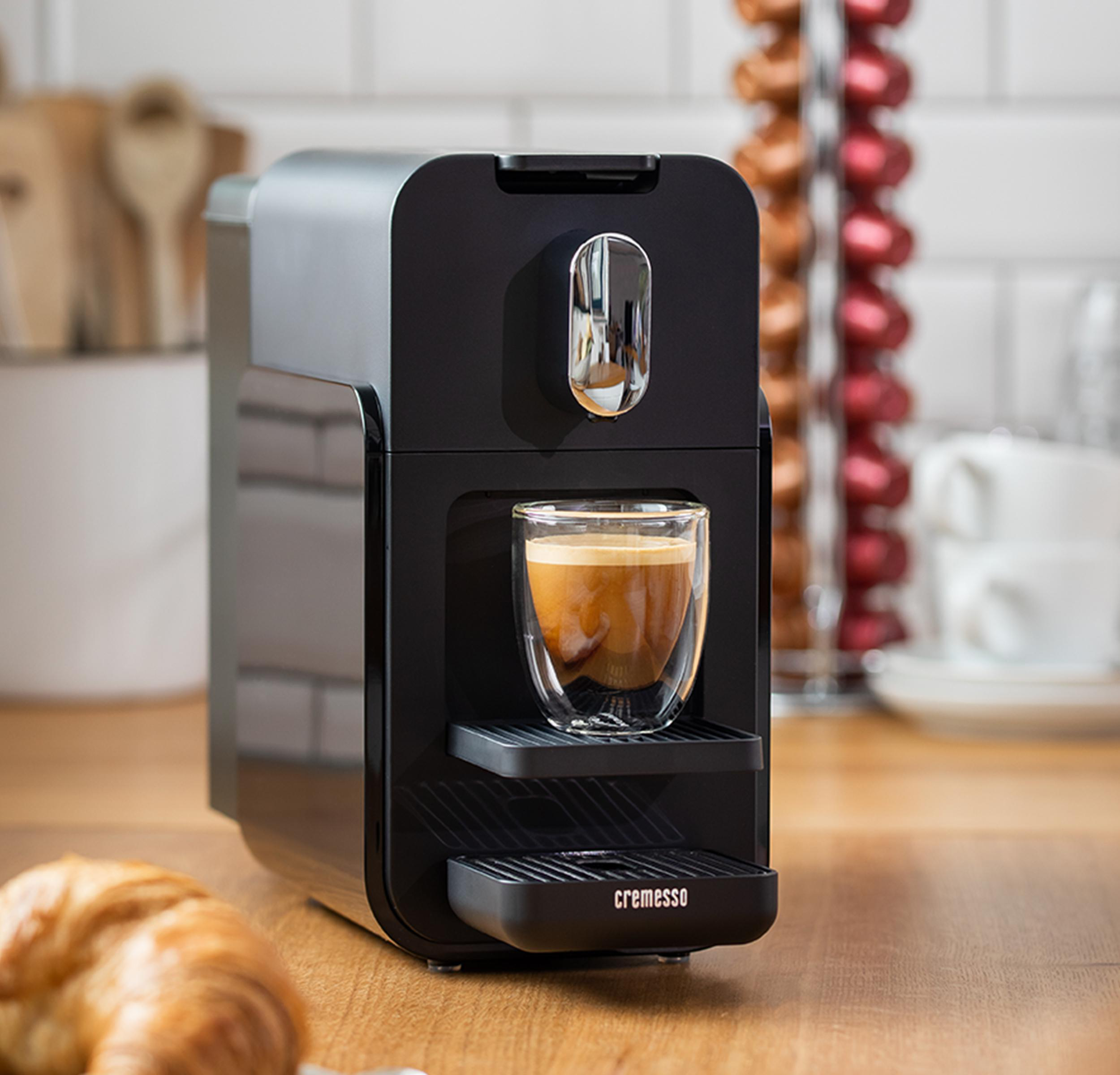 Schwarze Kaffeemaschine mit Kaffeeglas und Croissant auf Holzoberfläche.