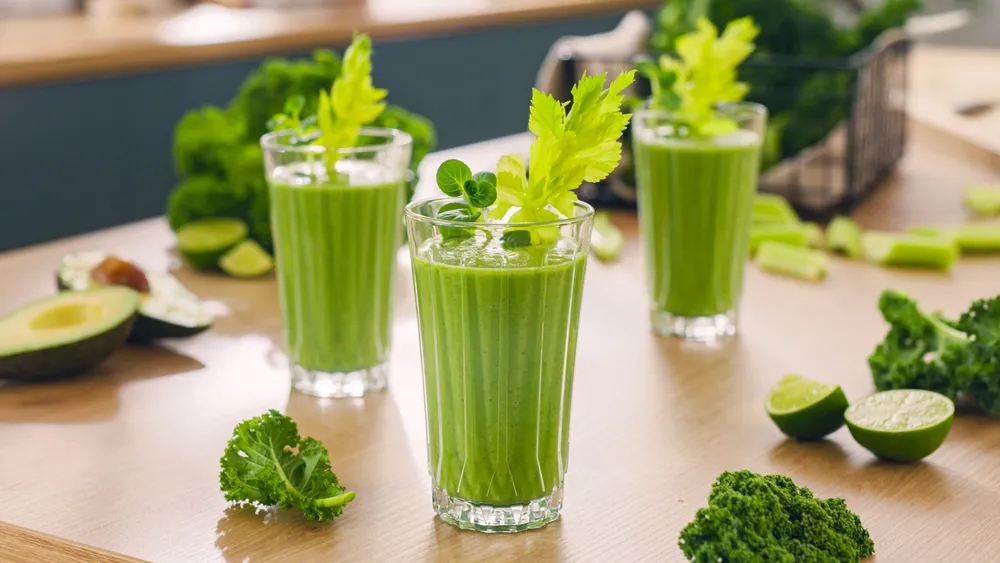 Három zöld smoothie zellerdíszítéssel, gyümölcsökkel egy fa felületen.