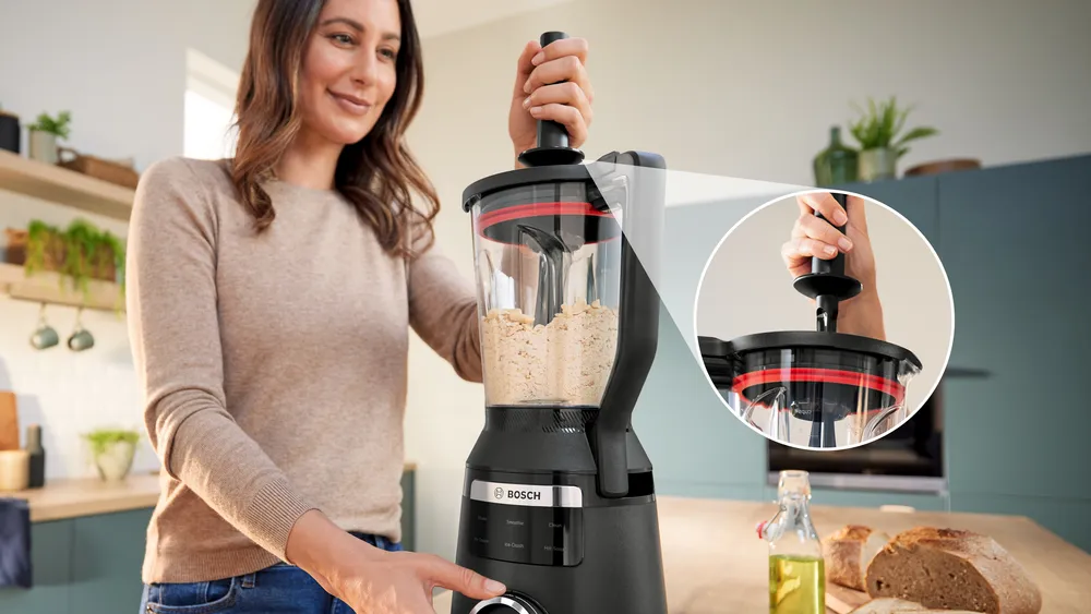 Nő egy fekete Bosch turmixgépet használ. A közelkép a mechanizmust mutatja. Kenyér és összetevők a háttérben.