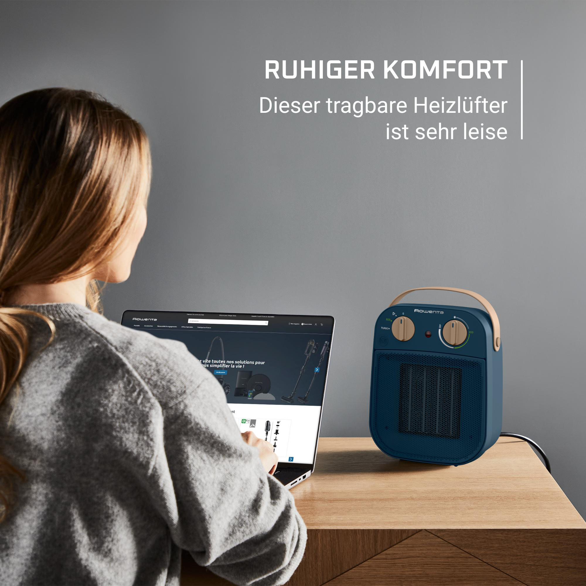 Frau am Laptop, neben blauem Heizlüfter. Text: 'RUHIGER KOMFORT'. Heizlüfter hat braunen Griff. Hintergrund: graue Wand.